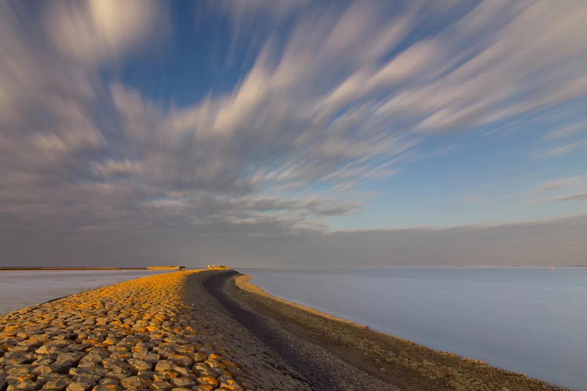 Ein schmaler Weg führt durch flaches Wasser auf den weiten Horizont zu, darüber ein ruhiger Himmel mit sanften Wolken.